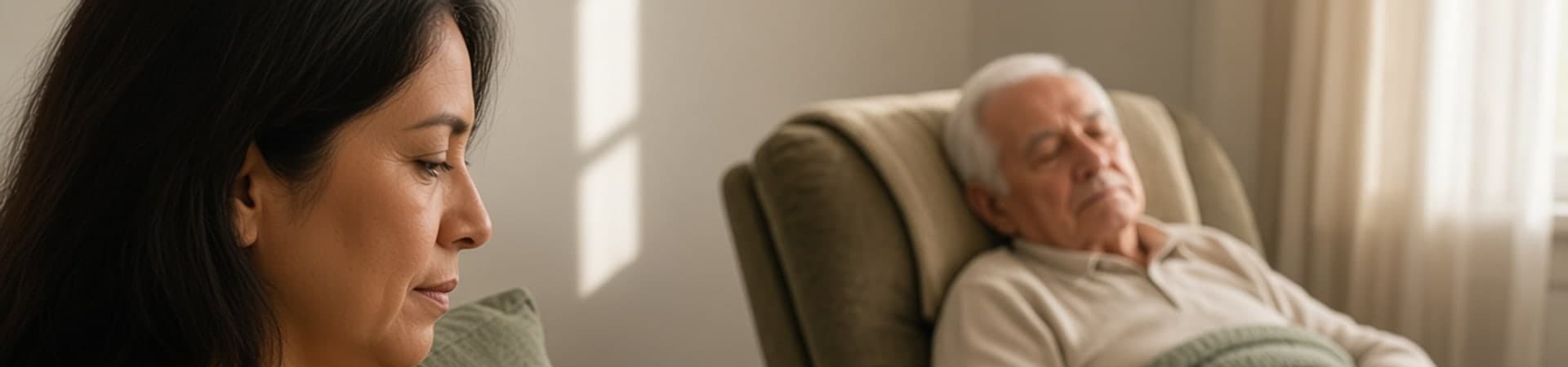A family caregiver sits quietly beside the older adult she cares for, who is resting in a recliner in a sunlit living room.